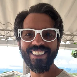Un hombre con barba y gafas blancas, sonriendo, en una terraza con montañas al fondo.