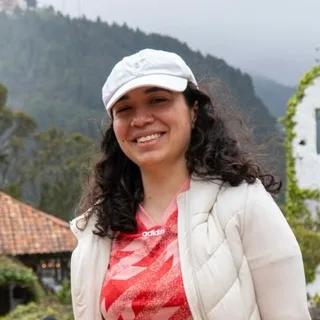 Una mujer de pelo rizado y gorra blanca, sonriendo, con montañas entre la niebla al fondo.